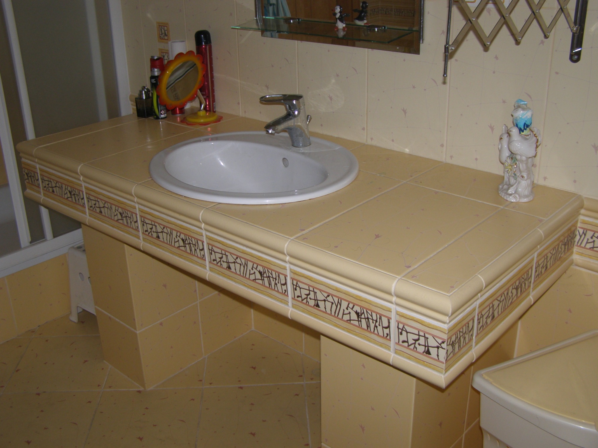 Beżowa umywalka wpuszczana w blat z płytek ceramicznych z dekoracyjnym paskiem, bateria chromowana, na blacie lusterko i kosmetyki, w tle płytki ścienne w delikatny wzór.