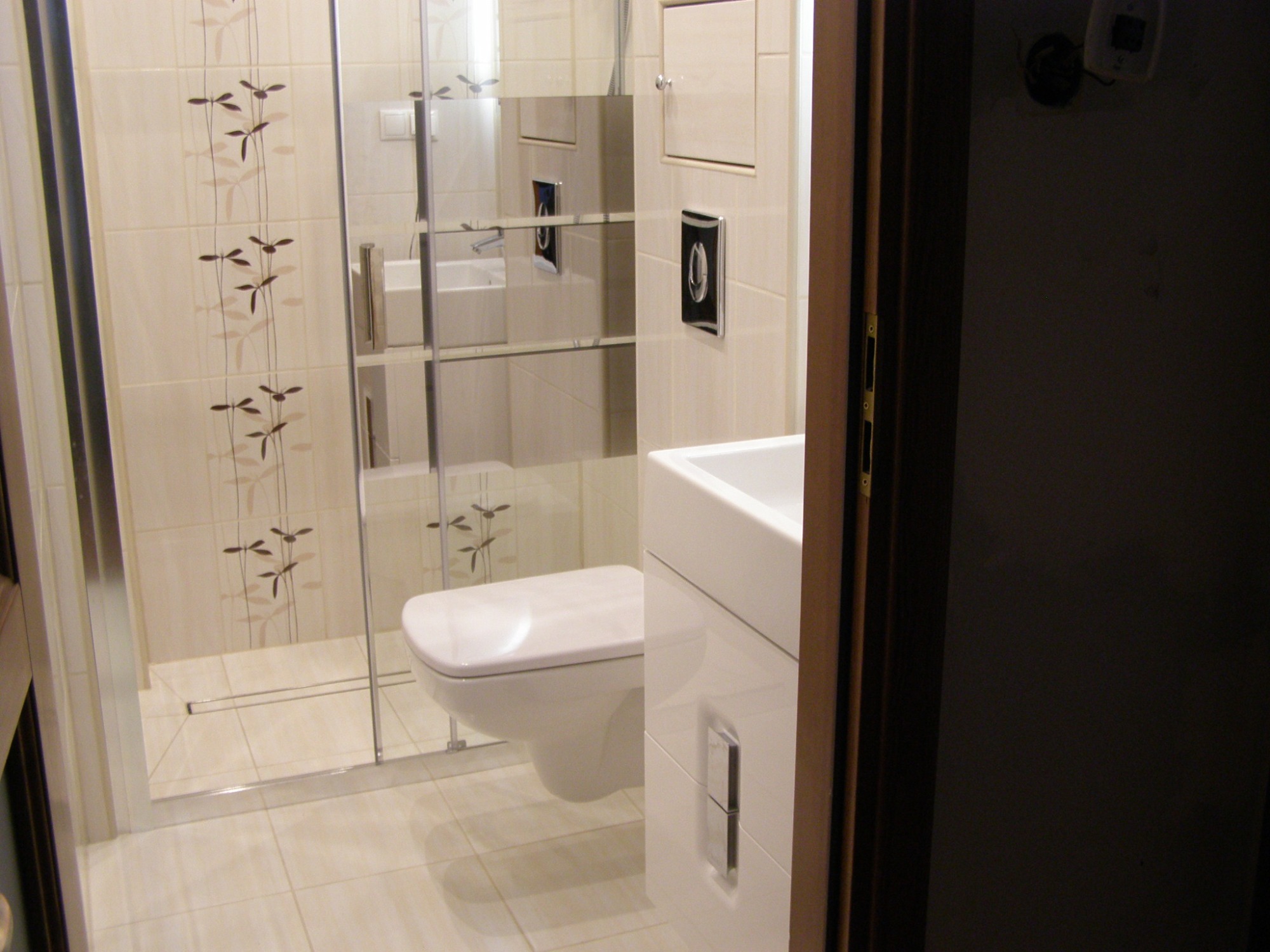 Nowoczesna łazienka z beżowymi płytkami w kwiatowy wzór, kabina prysznicowa typu walk-in z odpływem liniowym, podwieszana toaleta i umywalka nablatowa z szafką, widoczne zza uchylonych drzwi.