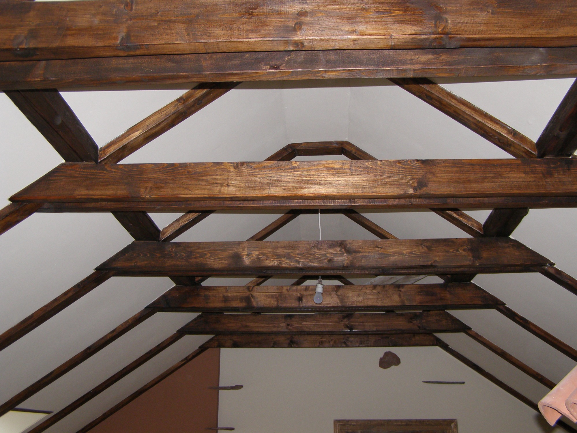Drewniana konstrukcja dachu z belek w ciemnym odcieniu brązu, widoczna od wewnątrz, surowy stan, żarówka na kablu zwisająca z sufitu, ściany w odcieniach bieli i beżu.