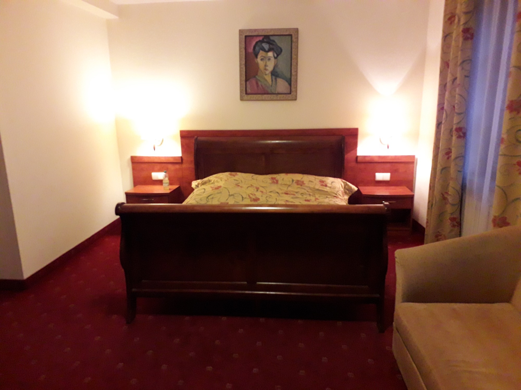 Pokój hotelowy z ciemnoczerwonym dywanem, drewnianym łóżkiem z zagłówkiem, szafkami nocnymi z lampkami, obrazem nad łóżkiem i beżowym fotelem w rogu