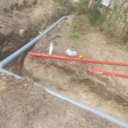 Rów w ziemi z ułożonymi szarymi rurami PCV, poziomica, kielnia, szpachelka i butelka środka czyszczącego leżące na ziemi, w tle roślinność.