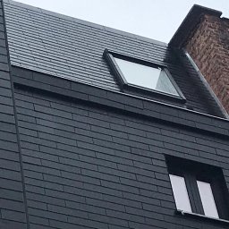 CM INVEST SPÓŁKA Z OGRANICZONĄ ODPOWIEDZIALNOŚCIĄ - Ciemna elewacja budynku z oknami i dachem pokrytym szarymi płytkami, widok z dołu, fragment sąsiedniego budynku z cegły w tle.