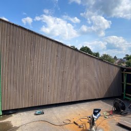 CM INVEST SPÓŁKA Z OGRANICZONĄ ODPOWIEDZIALNOŚCIĄ - Nowoczesna elewacja z pionowych desek w odcieniach brązu, widoczna z dołu pod kątem. Na pierwszym planie narzędzia pracy i resztki materiału. Błękitne niebo z chmurami w tle.