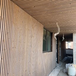 CM INVEST SPÓŁKA Z OGRANICZONĄ ODPOWIEDZIALNOŚCIĄ - Drewniana elewacja budynku z listew, widoczne okno i fragment sufitu z tego samego materiału. W tle elementy budowlane i narzędzia.