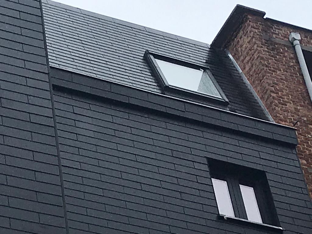 Ciemna elewacja budynku z oknami i dachem pokrytym szarymi płytkami, widok z dołu, fragment sąsiedniego budynku z cegły w tle.