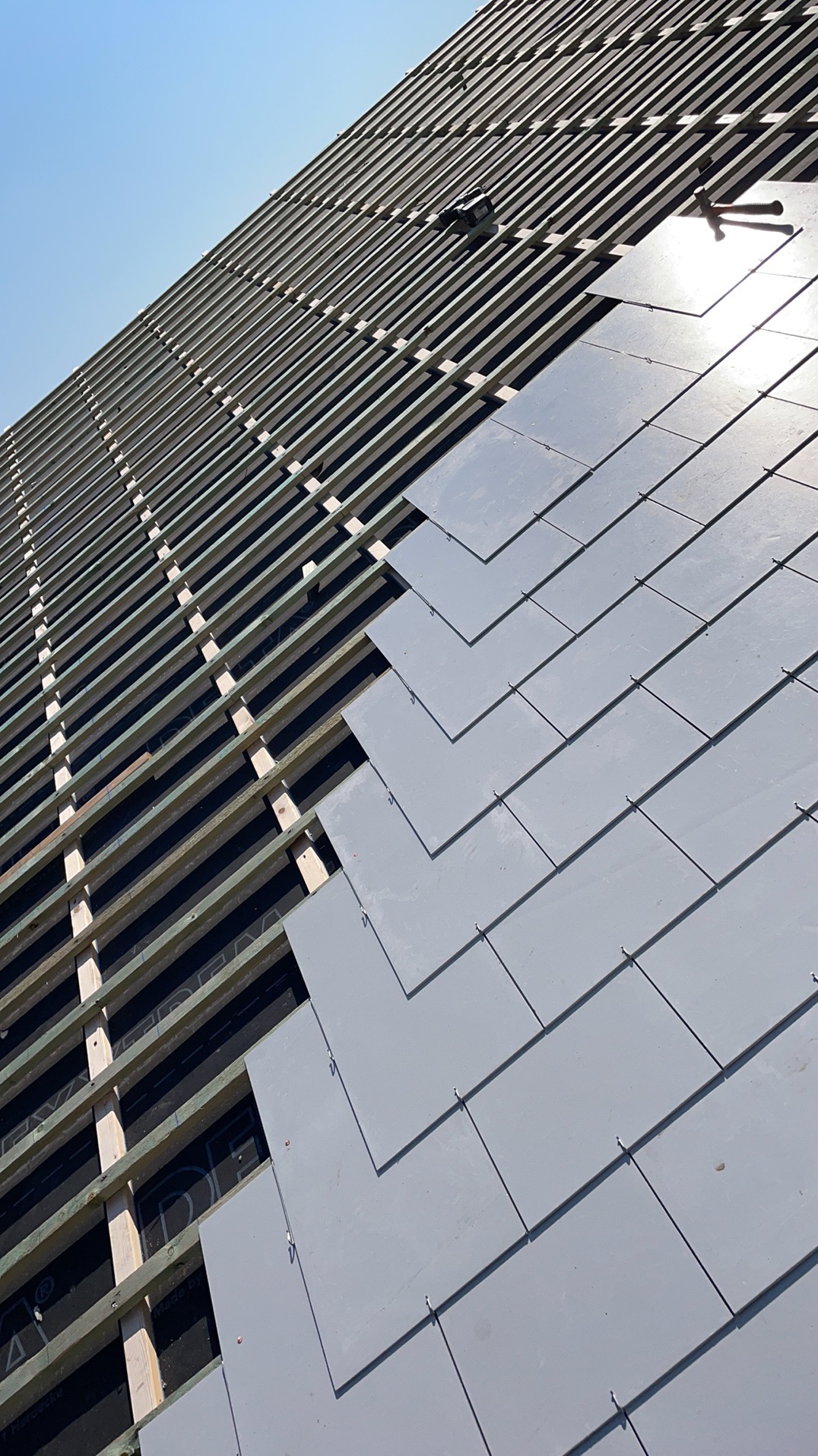 Montaż szarej dachówki na łatach z widoczną membraną. Częściowo pokryty dach w ukośnym ujęciu, narzędzia dekarskie na dachu.