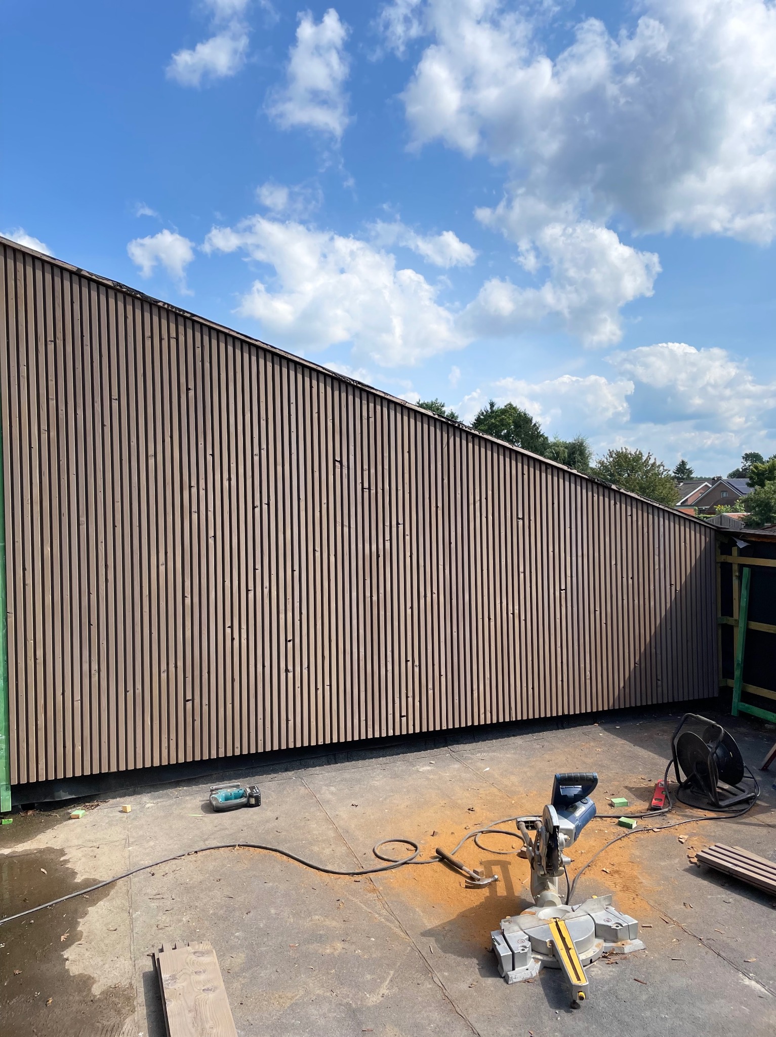 Nowoczesna elewacja z pionowych desek w odcieniach brązu, widoczna z dołu pod kątem. Na pierwszym planie narzędzia pracy i resztki materiału. Błękitne niebo z chmurami w tle.