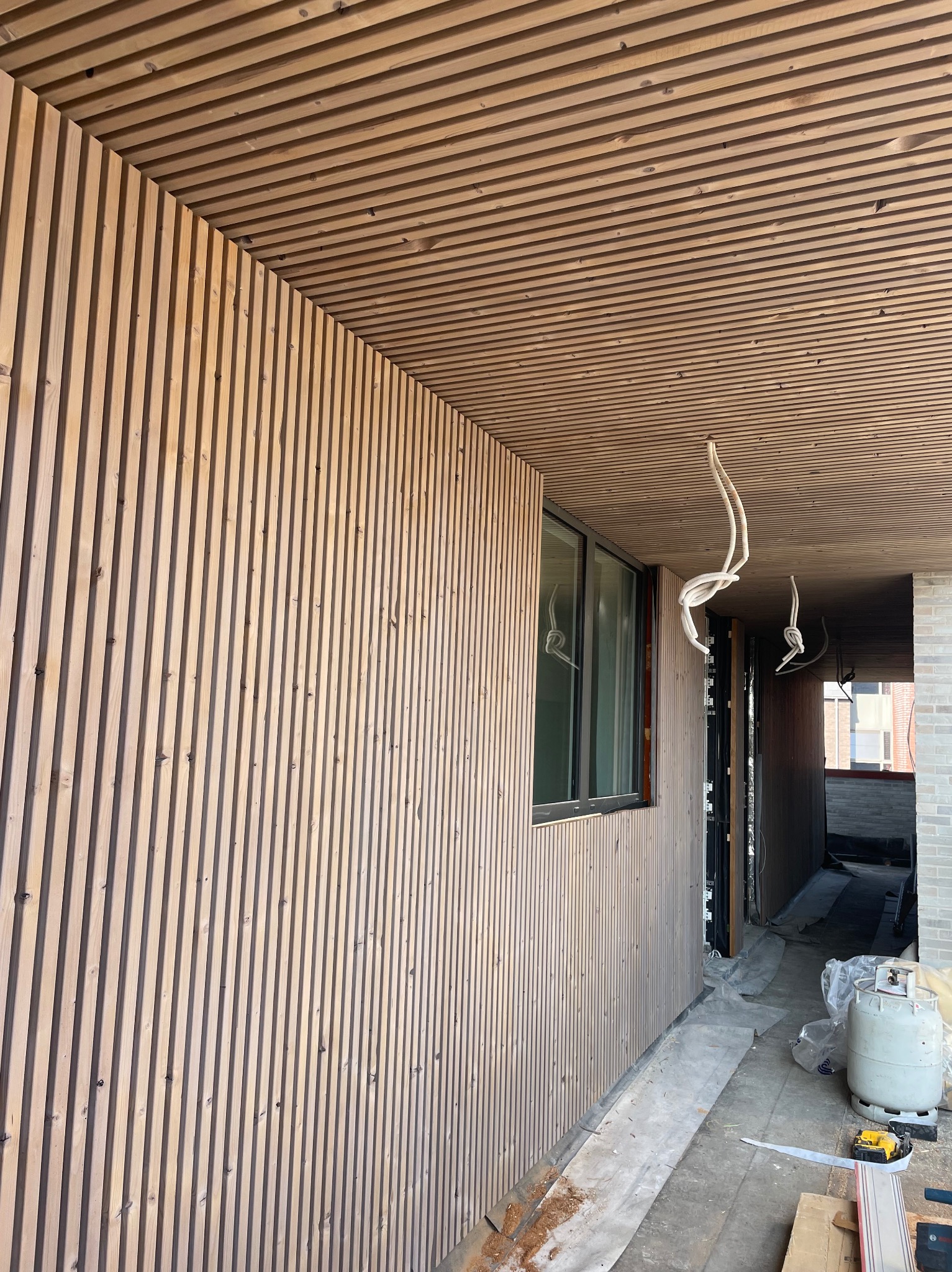 Drewniana elewacja budynku z listew, widoczne okno i fragment sufitu z tego samego materiału. W tle elementy budowlane i narzędzia.