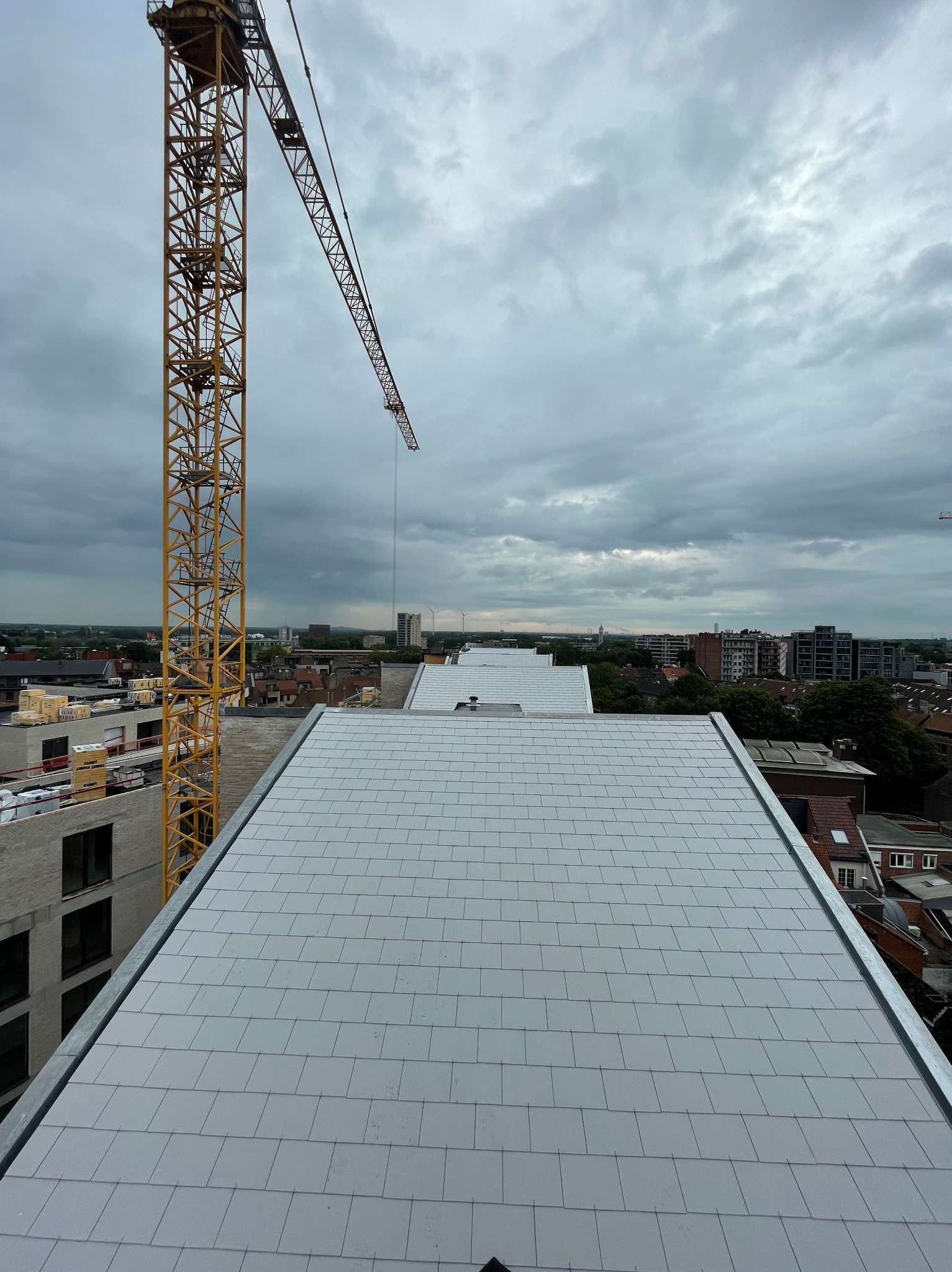 Nowy, jasnoszary dach z dachówki ceramicznej w miejskiej scenerii, z widocznym żurawiem budowlanym i budynkami w tle, uchwycony w pochmurny dzień.