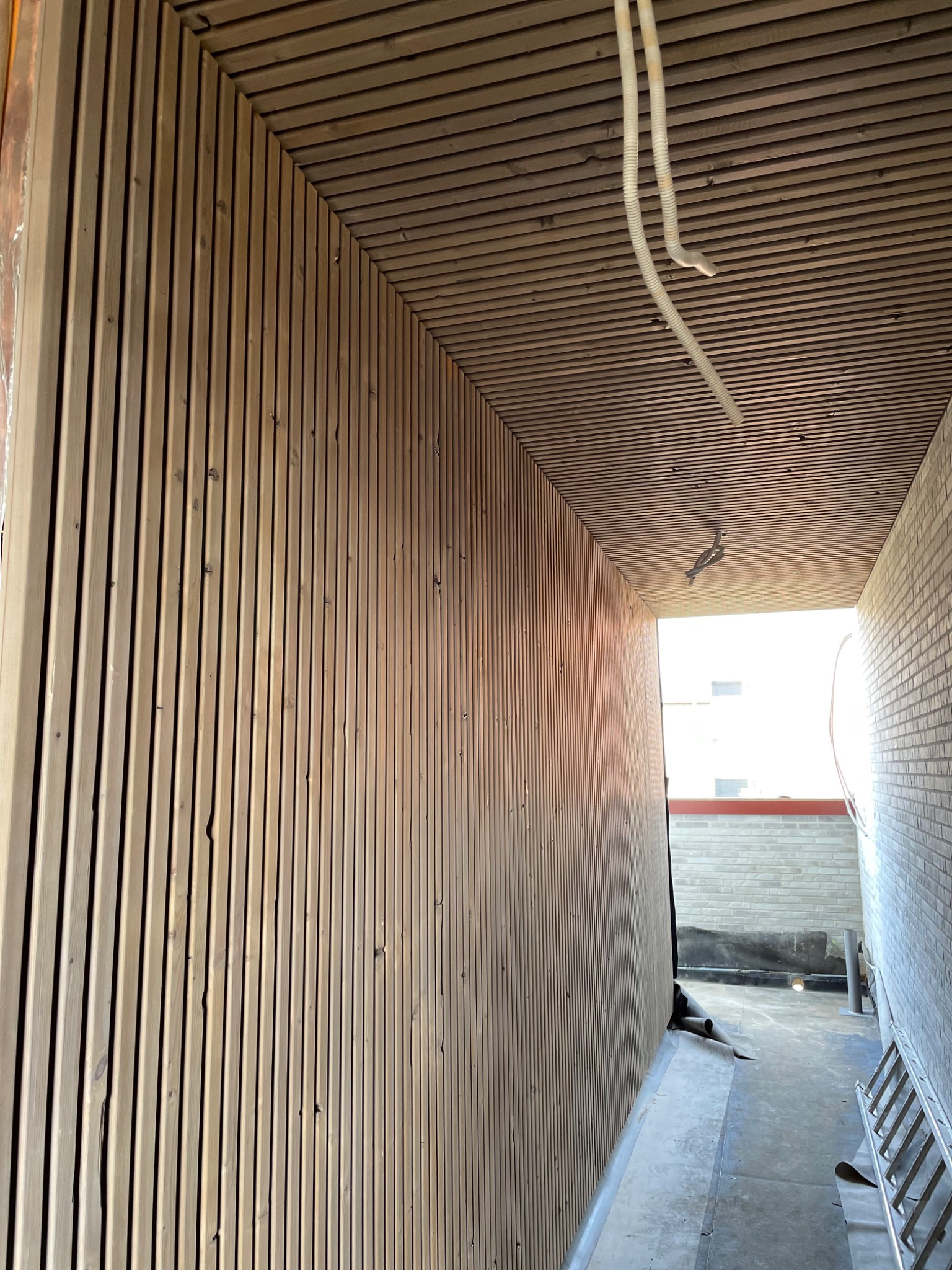 Wnętrze z drewnianą boazerią na ścianie i suficie, tworzącą korytarz. Widoczne przewody pod sufitem i drabina w tle. Ujęcie z perspektywy, podkreślające głębię przestrzeni.