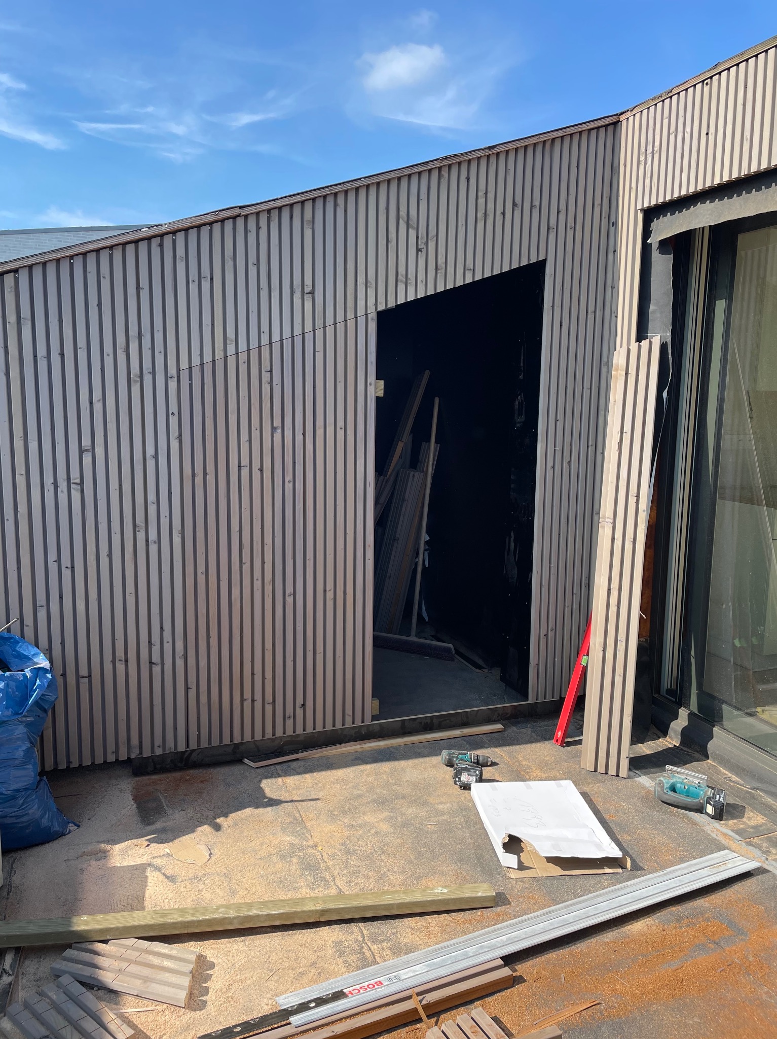 Montaż elewacji z drewnianych paneli pionowych. Widoczne narzędzia, fragment okna i niebieski worek. Prace w toku, surowe drewno i jasne oświetlenie.