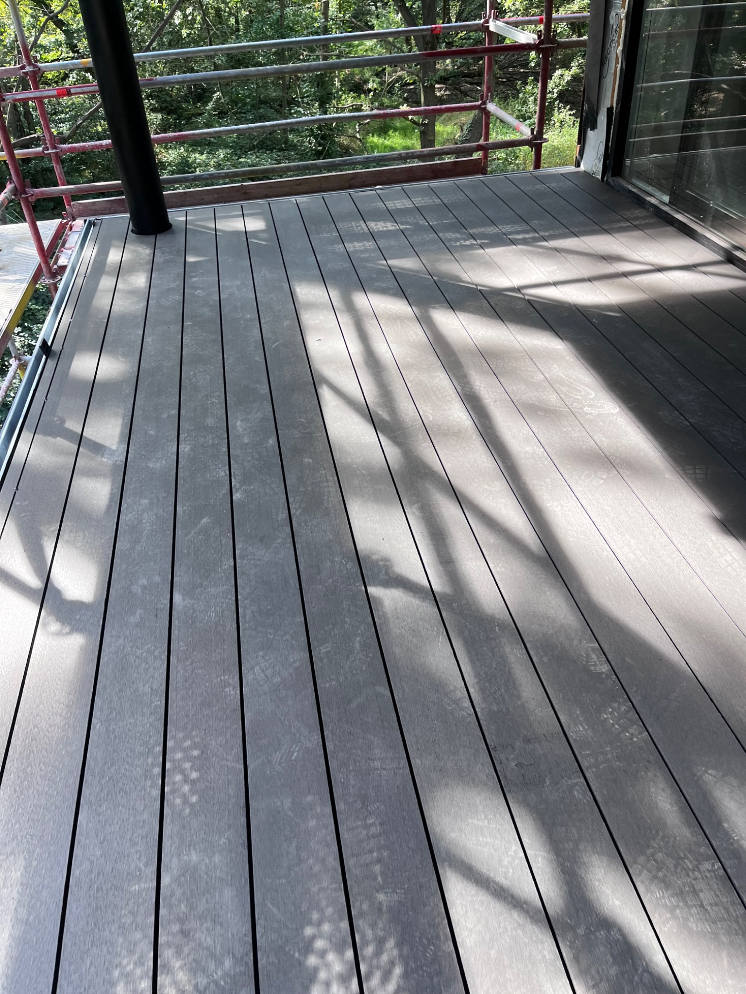 Nowoczesny taras kompozytowy w odcieniach szarości, z widocznymi cieniami drzew i fragmentem rusztowania. Minimalistyczny design, deski ułożone równolegle.