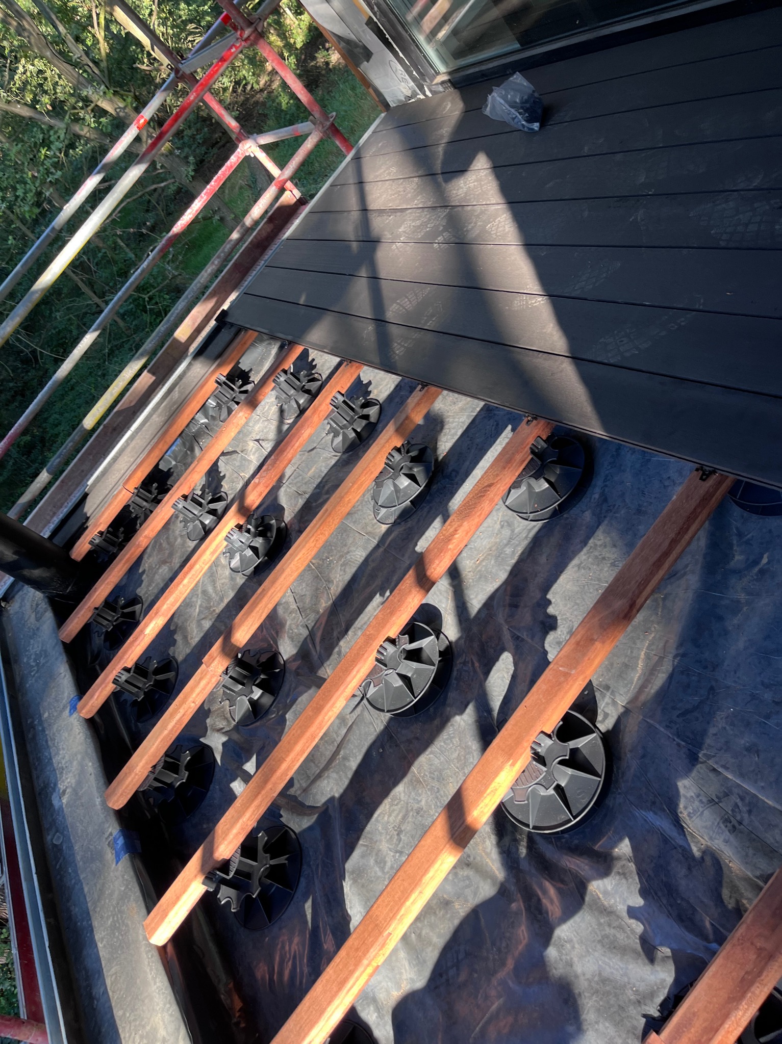 Montaż tarasu kompozytowego: legary na regulowanych wspornikach, częściowo ułożone deski. Widok z góry, z rusztowania. Czarna folia izolacyjna.