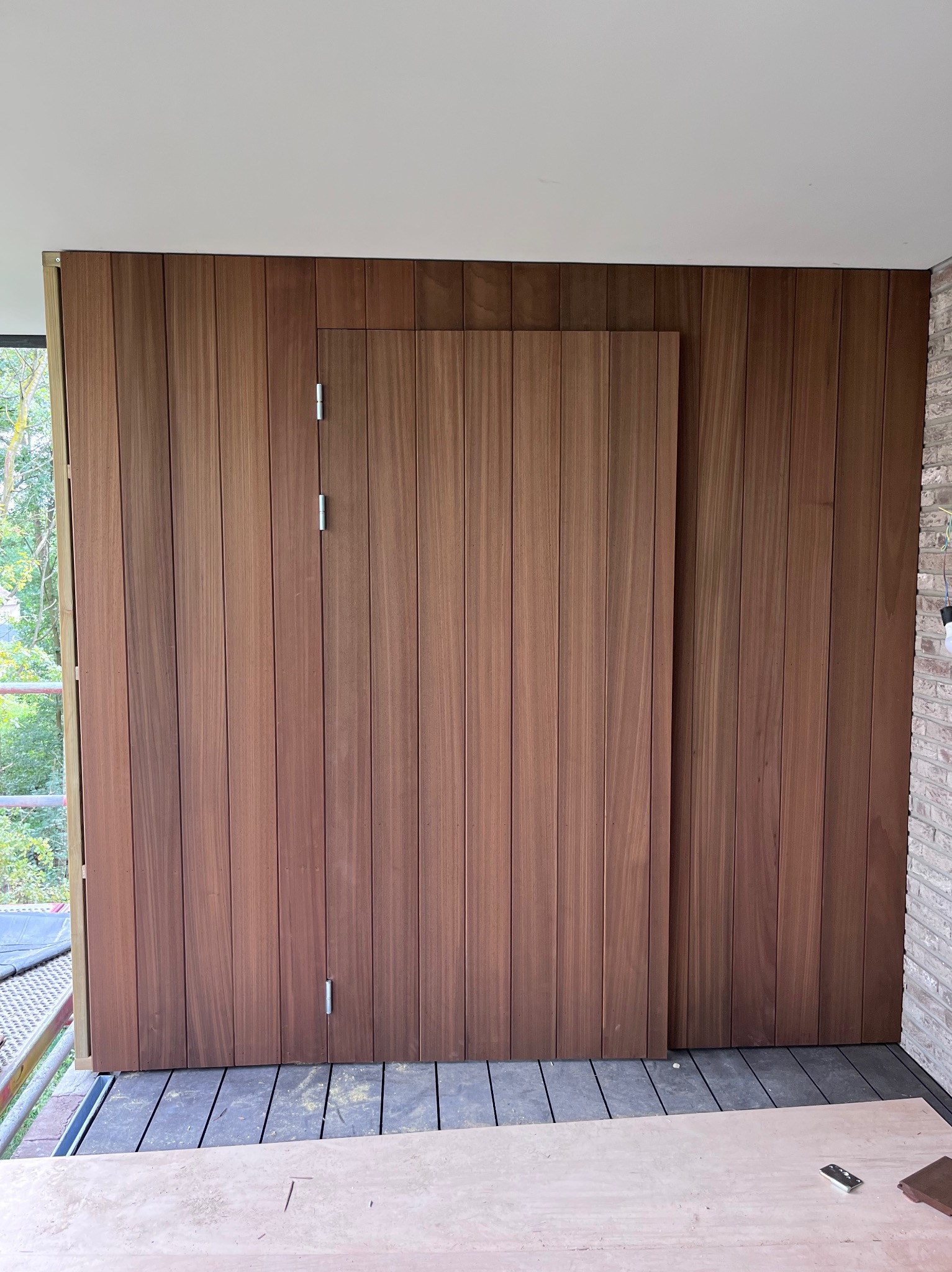 Drewniana ściana z ukrytymi drzwiami na tarasie, widok z bliska. Pionowe deski w ciepłym odcieniu brązu, fragment muru z cegły po prawej. Elementy rusztowania widoczne w tle.