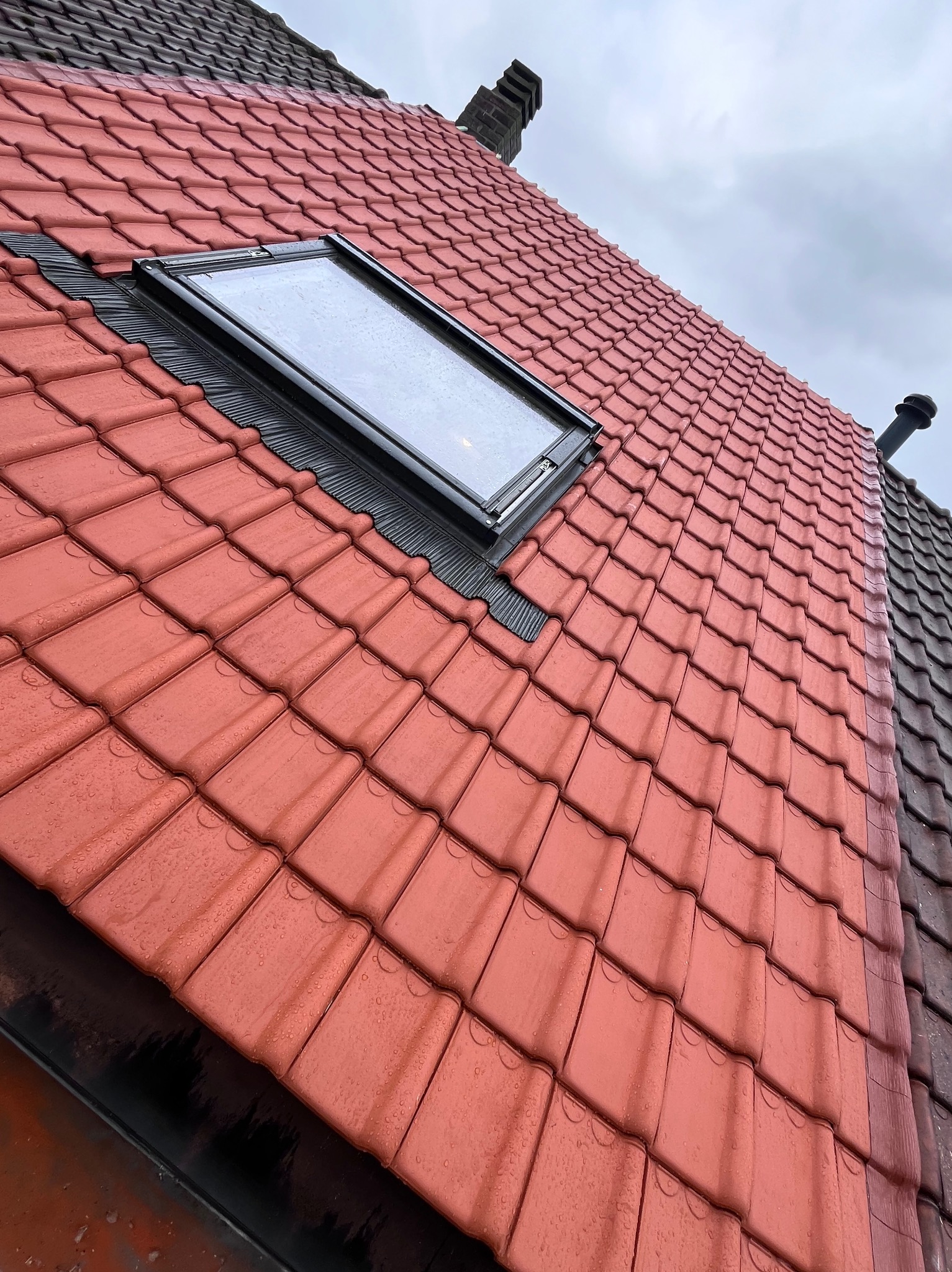 Czerwona dachówka ceramiczna z oknem dachowym, widok pod kątem. Na dachówce widoczne krople wody. Fragment dachu sąsiedniego budynku w odcieniach szarości.