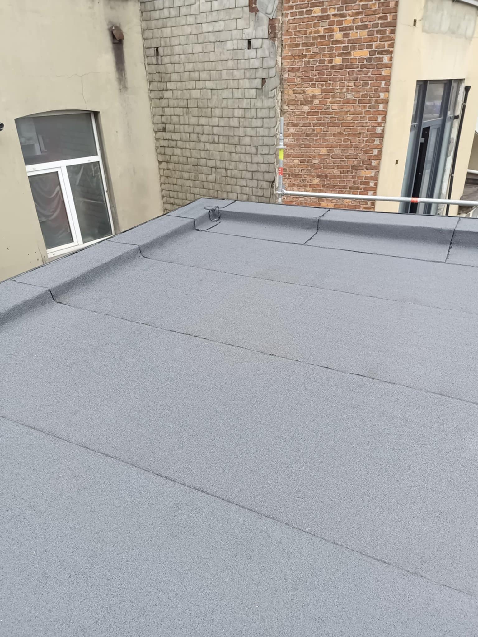 Szary, płaski dach pokryty papą termozgrzewalną, widok z góry. W tle budynki z cegły i okno. Minimalistyczne ujęcie detalu dachu po renowacji.