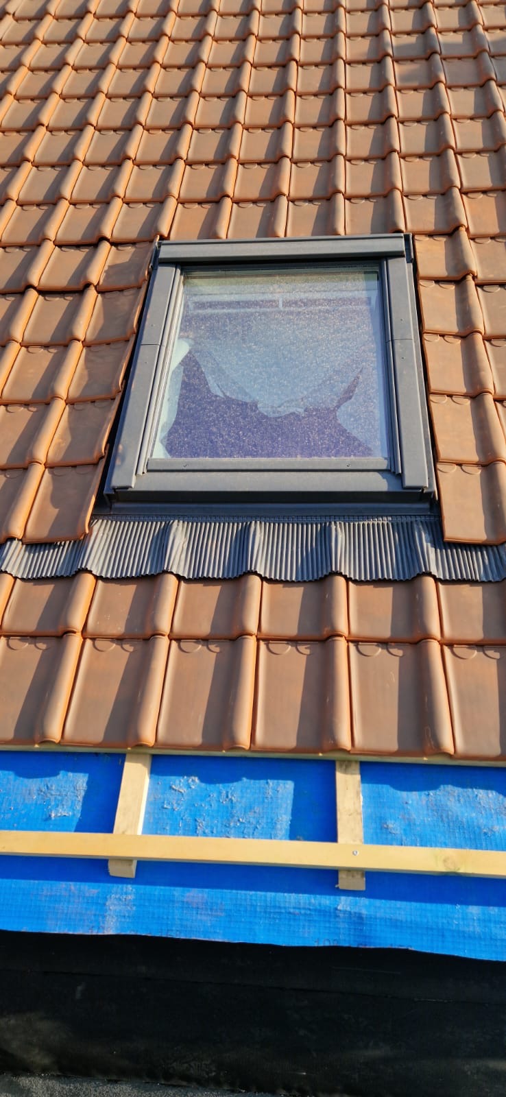 Montaż okna dachowego w brązowej dachówce ceramicznej. Widoczne elementy hydroizolacji i mocowania. Ujęcie z góry, w słoneczny dzień.