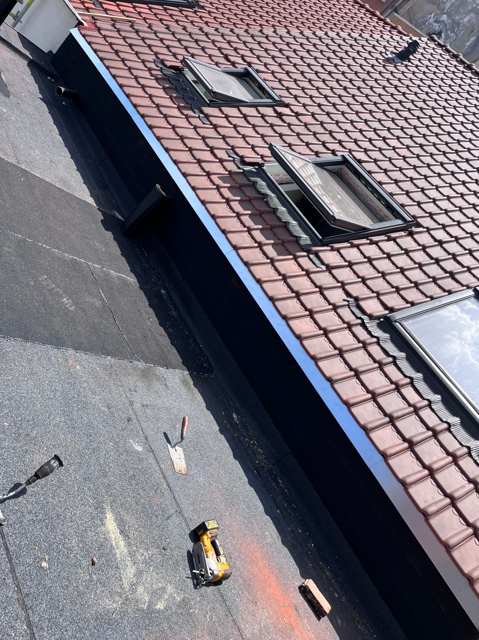 Czerwona dachówka z oknami dachowymi i metalowym obramowaniem. Widoczne narzędzia dekarskie na czarnej papie. Ujęcie z góry, ostre światło.