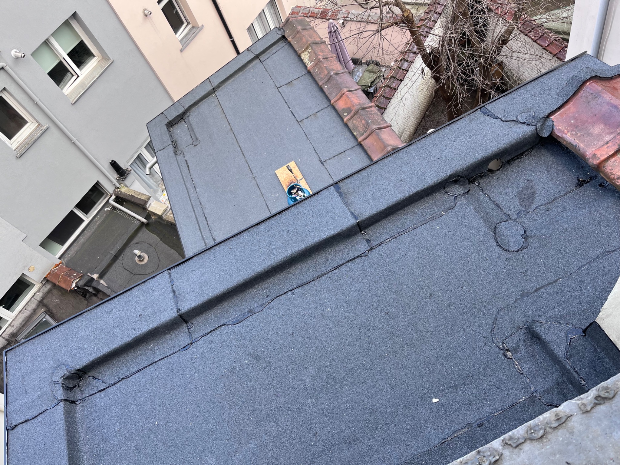 Płaski dach pokryty papą z widocznymi łączeniami i śladami napraw. W tle budynki mieszkalne i fragment drzewa bez liści. Na dachu leży deska z narzędziami.