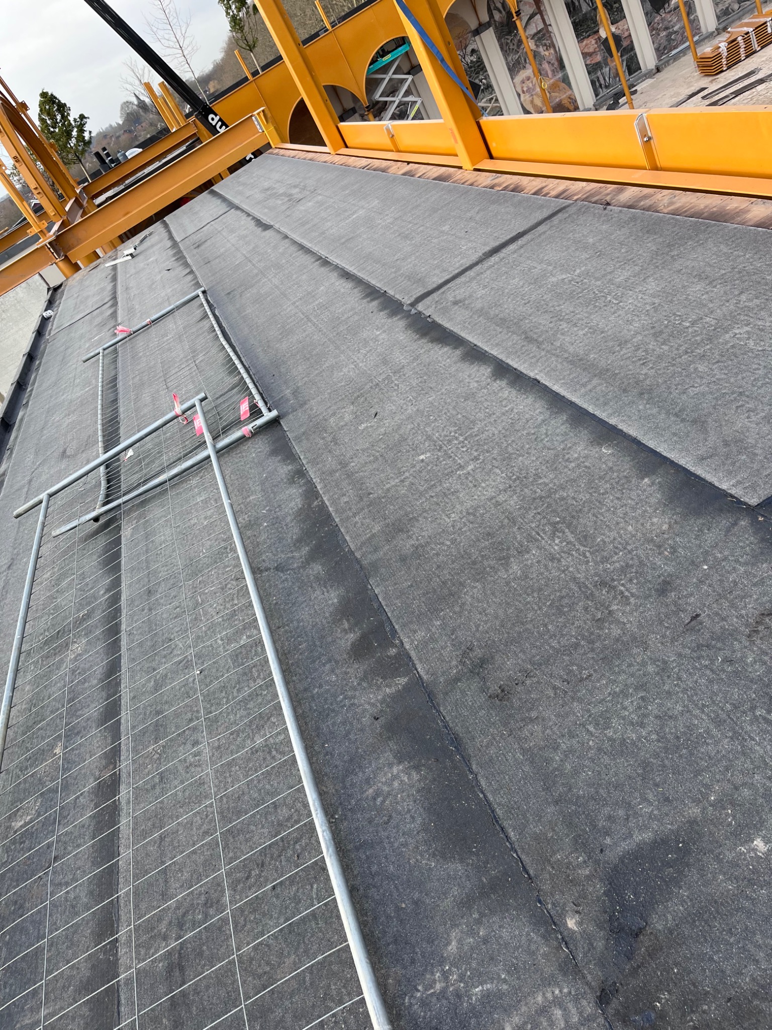Czarny, płaski dach pokryty papą, zabezpieczony metalowymi barierkami i elementami rusztowania w kolorze żółtym. Widok z góry, ukośny kadr.