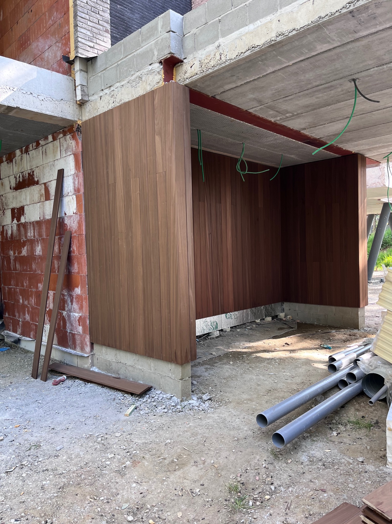 Montaż drewnianej elewacji na surowej konstrukcji budynku. Widoczne deski elewacyjne, betonowe fundamenty i instalacje. Prace w toku, surowy stan wykończenia.