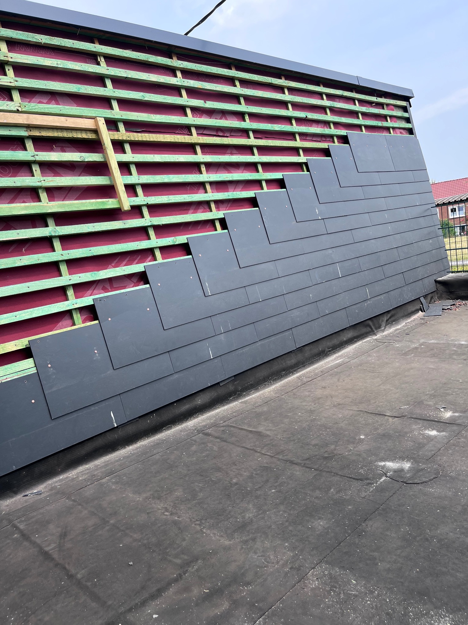 Układanie grafitowych płyt na dachu z widoczną konstrukcją drewnianą i membraną. Częściowo pokryty dach w trakcie prac dekarskich, widok z góry na płaską powierzchnię dachu.