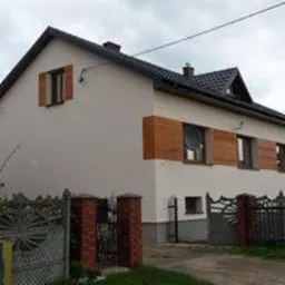 Dwukondygnacyjny dom z beżową elewacją, ozdobiony drewnianymi panelami imitującymi okiennice, widoczne metalowe ogrodzenie z ceglanymi słupkami, pochmurne niebo.