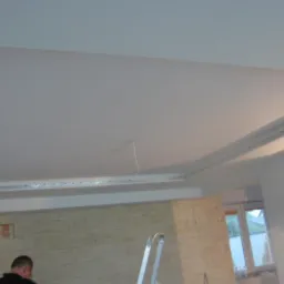 Wykończony sufit podwieszany z oświetleniem LED, widoczny fragment ściany z kamienia dekoracyjnego i drabina aluminiowa w pomieszczeniu.