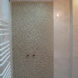 Łazienka w trakcie wykańczania: mozaika z drobnych płytek w odcieniach szarości i beżu, jasne płytki imitujące marmur, biały grzejnik drabinkowy, widoczne przyłącza wodne.