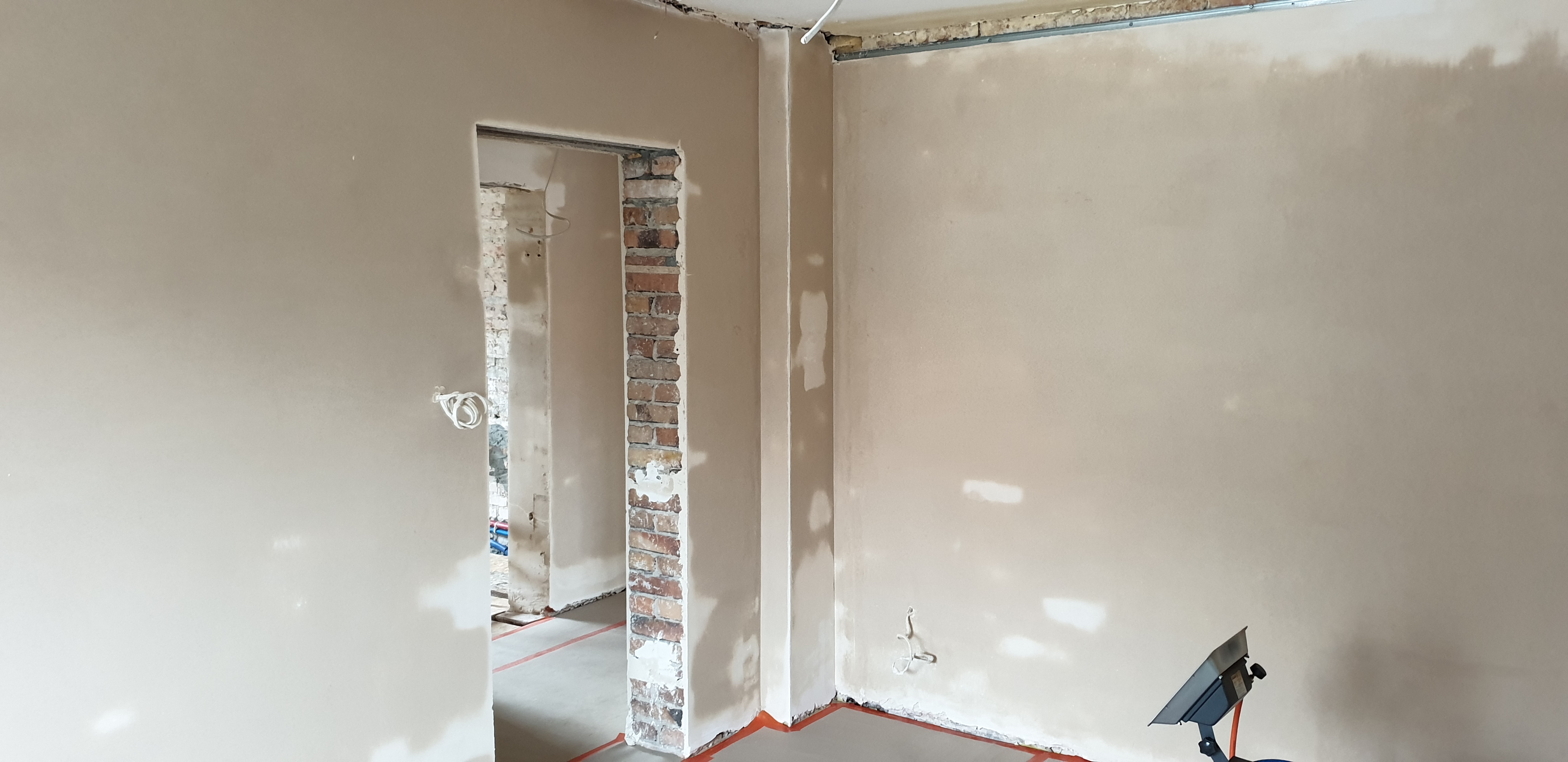 Wnętrze w trakcie remontu: ściany po szpachlowaniu, widoczne fragmenty cegieł przy otworze drzwiowym, zabezpieczona podłoga folią, lampa halogenowa oświetlająca ścianę.