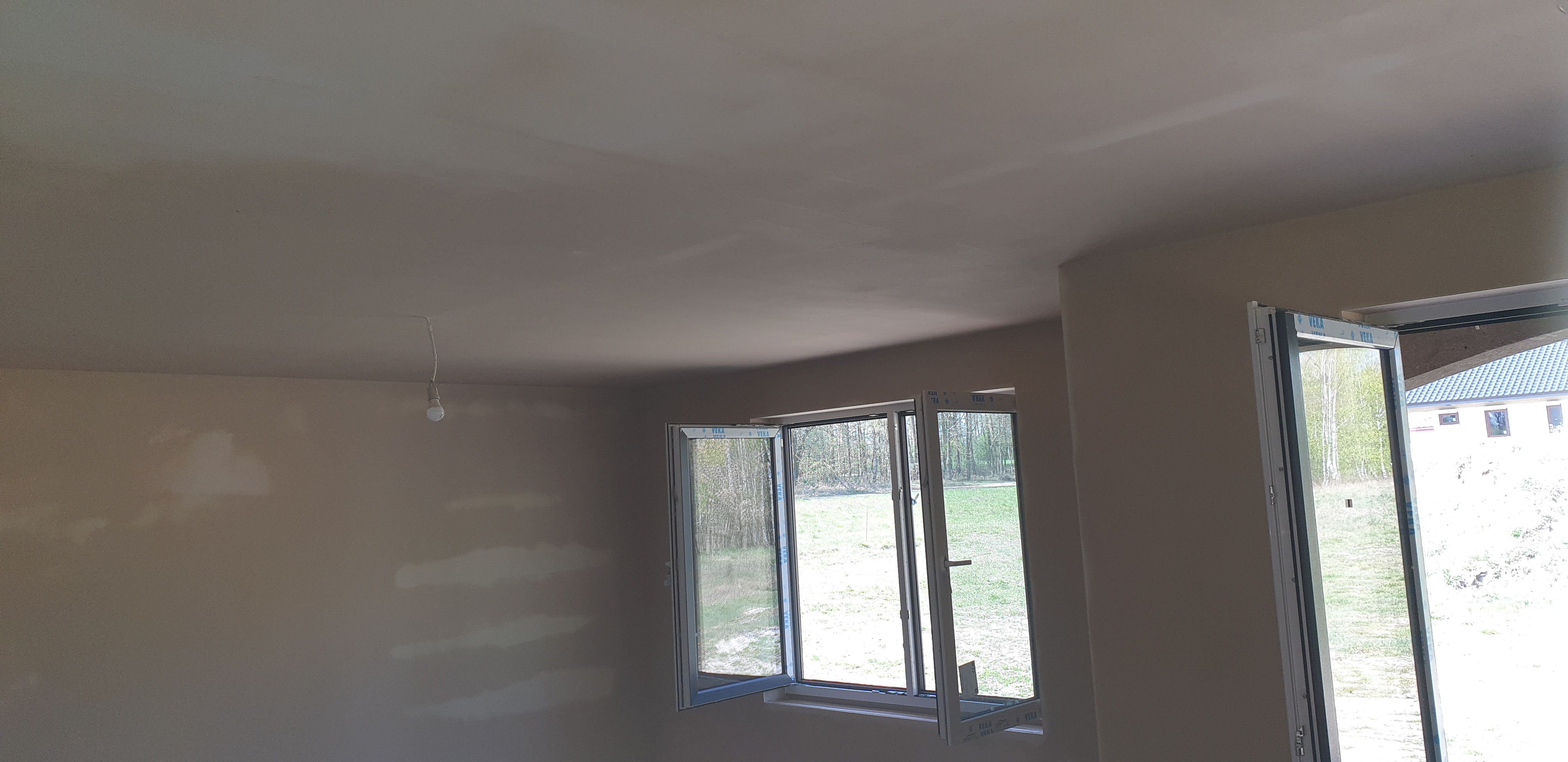 Wnętrze w stanie deweloperskim z przygotowaną, gładką powierzchnią ścian i sufitu, widoczne dwa okna z białymi ramami, jedno otwarte, surowa żarówka zwisająca z sufitu.