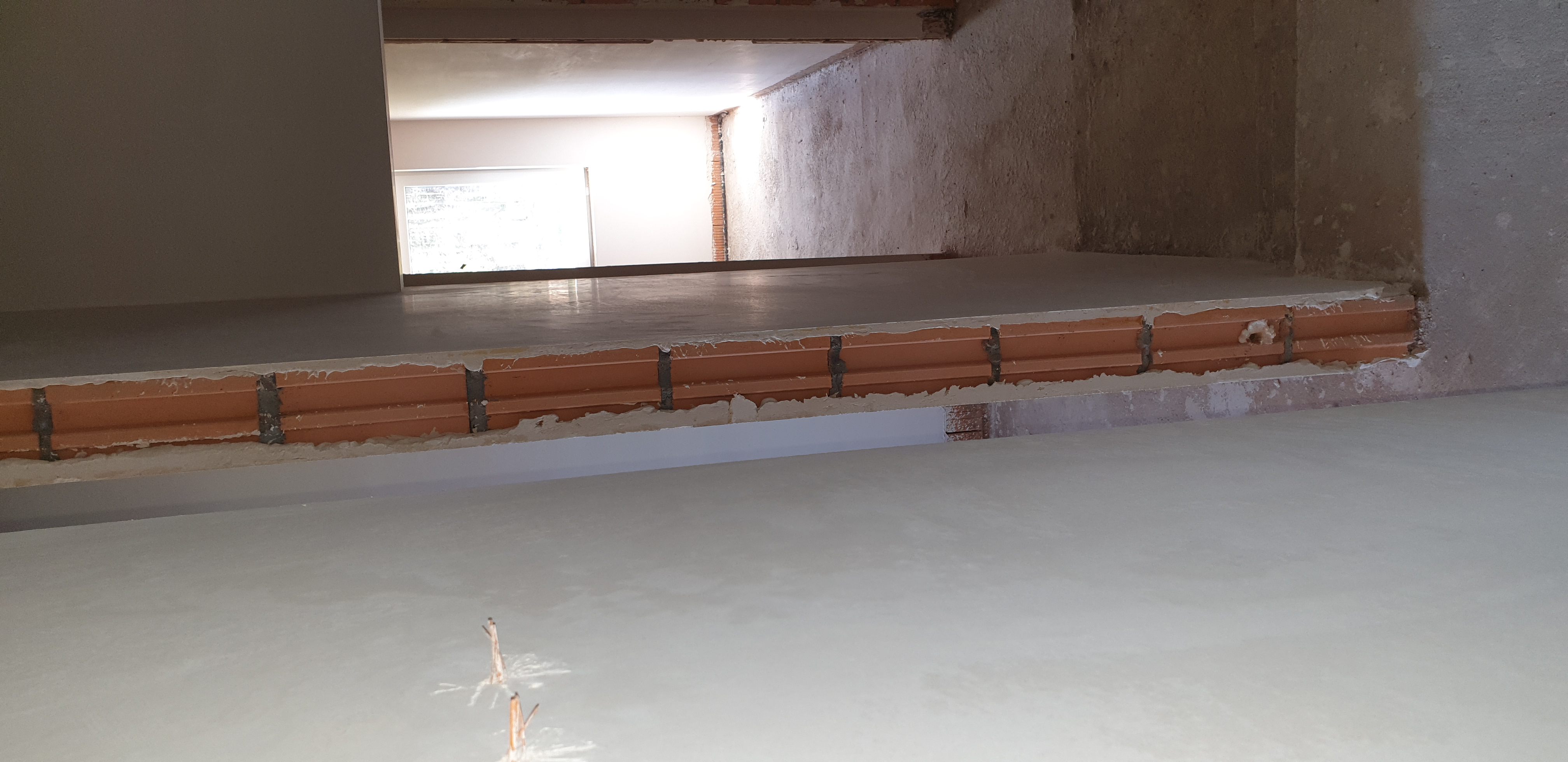 Surowy stan wnętrza w trakcie prac: widok na strop z ceglanym wypełnieniem i świeżo nałożoną, gładką warstwą, z fragmentem ściany w tle i oknem w oddali.