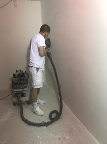 Pracownik szlifuje ścianę w rogu pomieszczenia za pomocą szlifierki z odkurzaczem przemysłowym, przygotowując powierzchnię pod dalsze prace wykończeniowe.