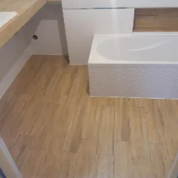 Nowoczesna łazienka z drewnopodobną podłogą, białą wanną z teksturowanym panelem i umywalką na drewnianym blacie.