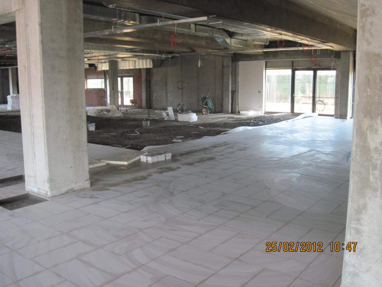 Układanie płytek podłogowych w przestronnym, surowym wnętrzu w Obrzycko. Widoczne betonowe kolumny i przygotowane podłoże pod dalsze prace wykończeniowe.