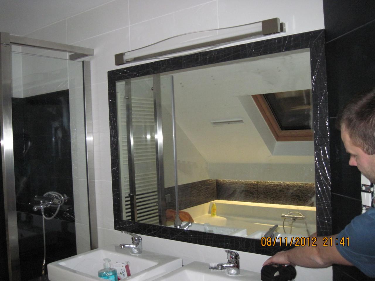 Wykończona łazienka z kabiną prysznicową i umywalką z lustrem w czarnej ramie. W lustrze odbija się wanna i okno dachowe. Widoczny fragment osoby.