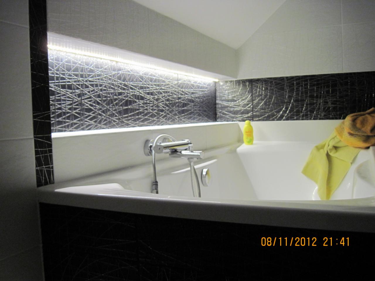 Wykończona łazienka z wanną, czarno-białe płytki z geometrycznym wzorem, oświetlenie LED nad wanną. Żółty ręcznik i butelka płynu do kąpieli na wannie.