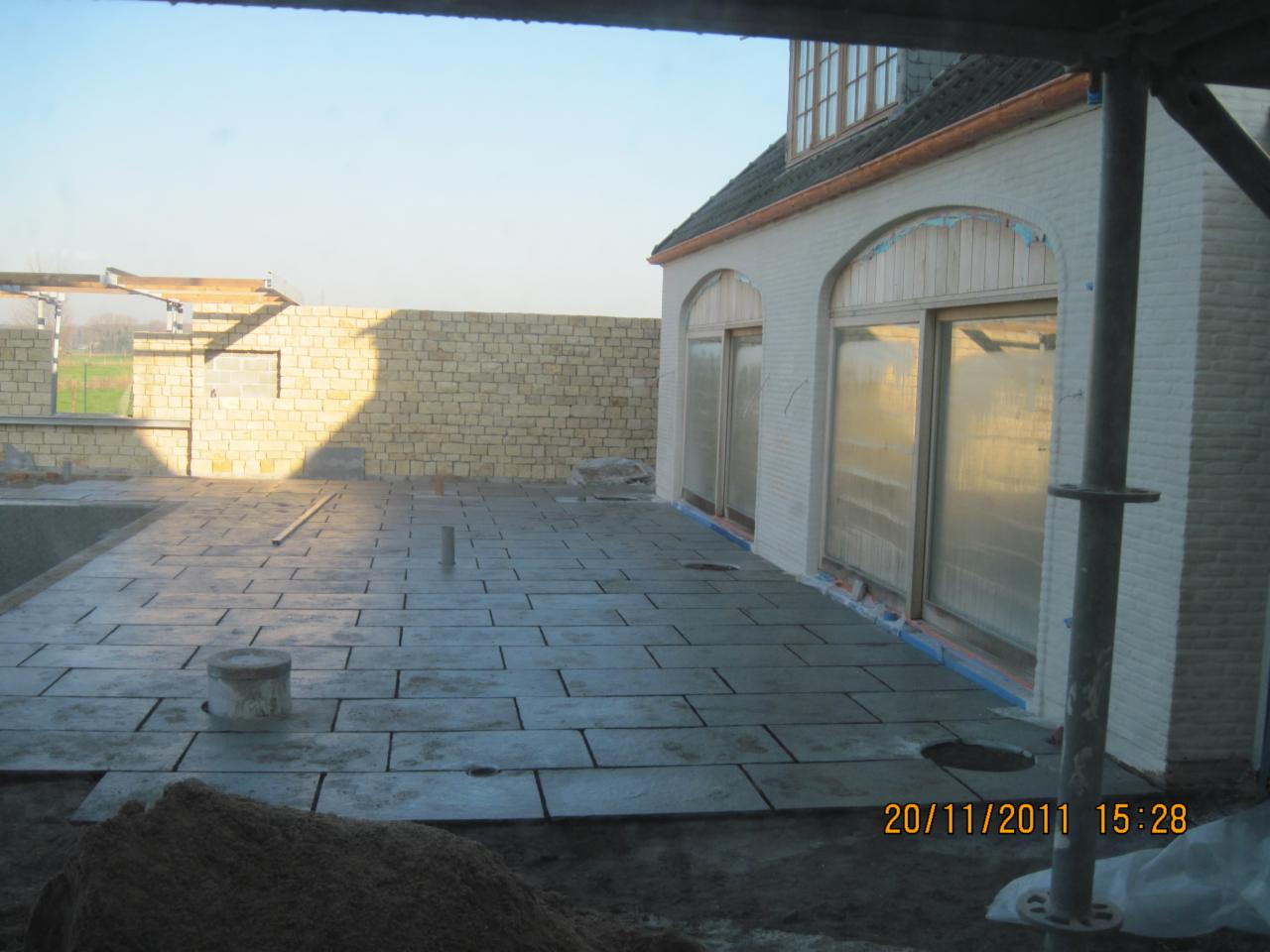 Układanie szarej kostki brukowej na tarasie domu z białej cegły. Widoczne okna zabezpieczone folią. Na pierwszym planie piasek i data wykonania zdjęcia.