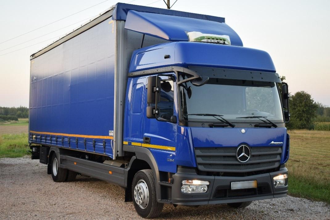 Niebieski samochód ciężarowy Mercedes Atego z plandeką na tle polnego krajobrazu o zmierzchu.
