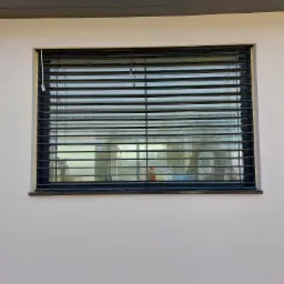 Zamknięte, poziome żaluzje fasadowe w oknie budynku z szarą elewacją i ciemnym podbiciem dachowym.