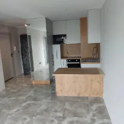 Nowoczesna kuchnia w trakcie wykończenia, widoczne szare płytki podłogowe, biało-drewniane szafki kuchenne, wyspa z drewnianym blatem i zabezpieczone folią drzwi w tle.