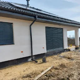 Nowoczesny dom jednorodzinny z zamontowanymi szarymi roletami zewnętrznymi na oknach, widoczne przewody elektryczne wystające ze ściany oraz teren wokół domu w trakcie prac wykończeniowych.