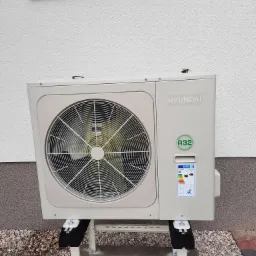 Zewnętrzna jednostka klimatyzacji Hyundai, umieszczona na metalowym stelażu, z widoczną etykietą energetyczną i oznaczeniem czynnika chłodniczego R32, na tle jasnej elewacji budynku i żwirowego...