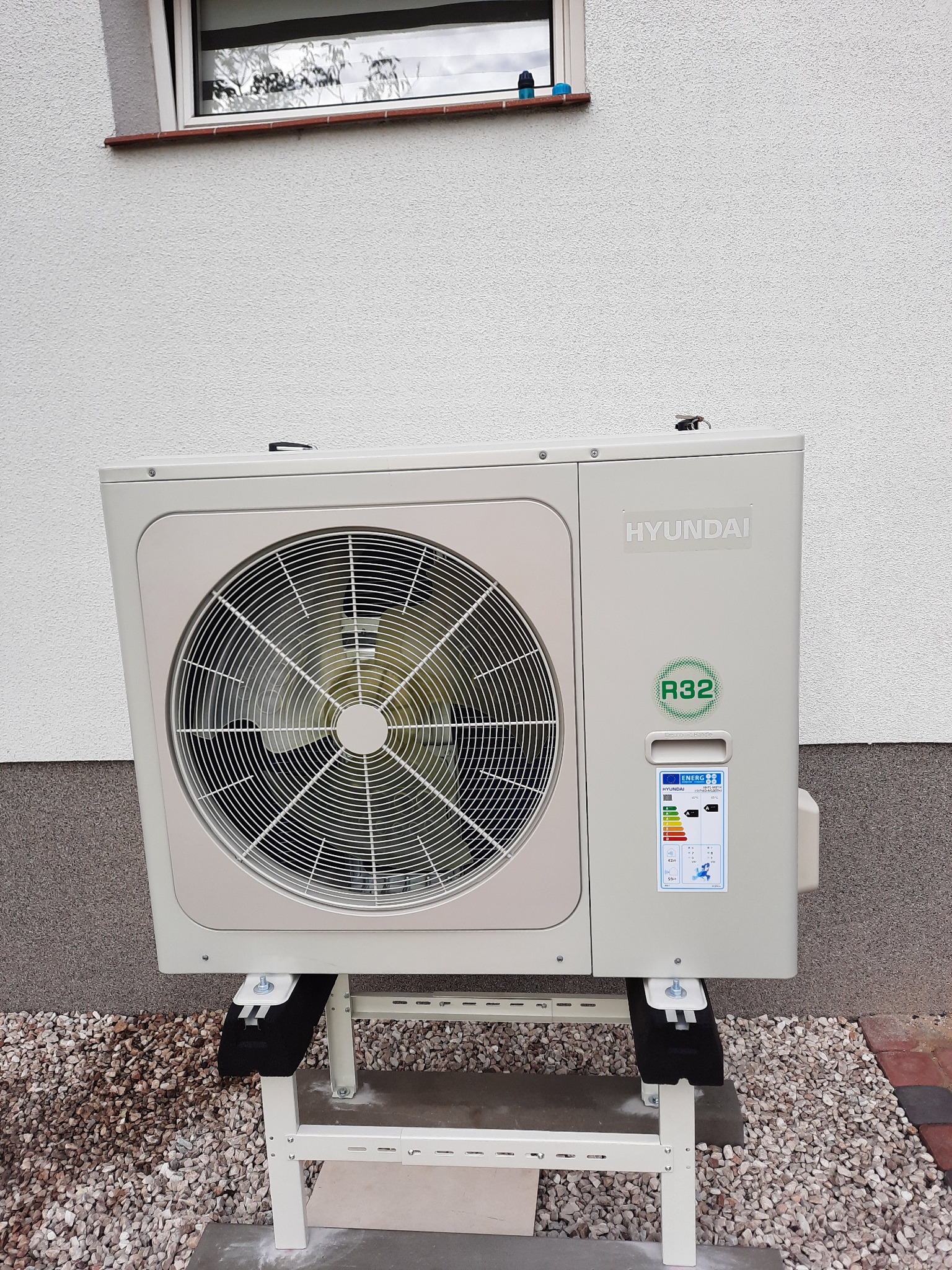 Zewnętrzna jednostka klimatyzacji Hyundai, umieszczona na metalowym stelażu, z widoczną etykietą energetyczną i oznaczeniem czynnika chłodniczego R32, na tle jasnej elewacji budynku i żwirowego...