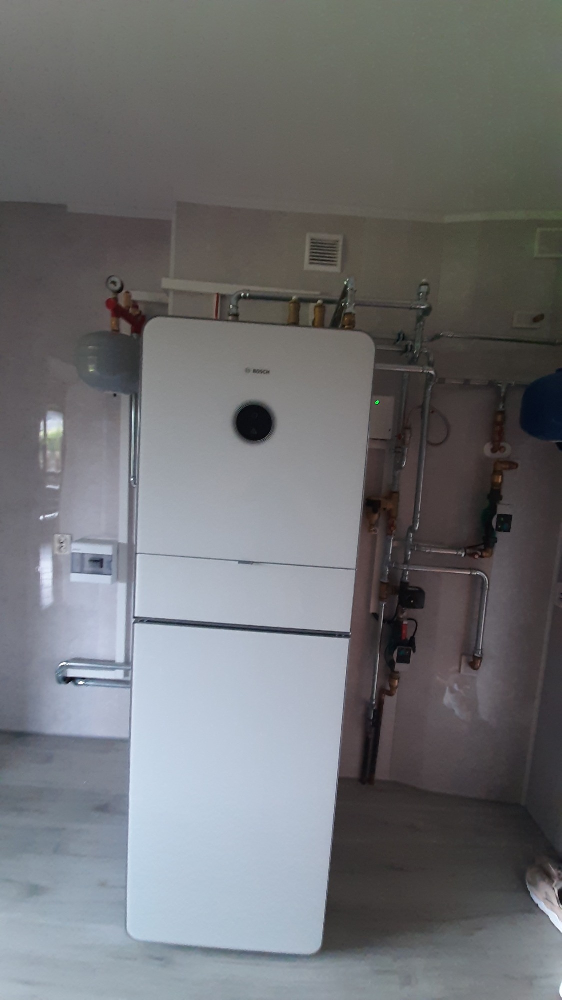 Biały, nowoczesny piec grzewczy marki Bosch z podłączeniami hydraulicznymi na ścianie w szarym pomieszczeniu. Widoczny zbiornik wyrównawczy i elementy instalacji.