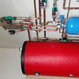 Czerwony zbiornik ciepłej wody użytkowej z niebieskim zbiornikiem wyrównawczym i miedzianą instalacją grzewczą na białej ścianie.