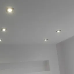 Biały sufit z sześcioma kwadratowymi, wpuszczanymi lampami halogenowymi, widoczny fragment okna z zasłoną i wnęka w ścianie.