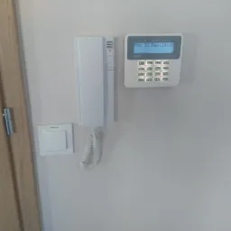 Biały domofon z panelem dotykowym Satel i słuchawką na ścianie, obok drzwi z drewnianą ościeżnicą i włącznika światła.