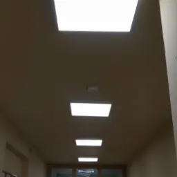 Korytarz oświetlony szeregiem kwadratowych paneli LED w suficie, widok od dołu w kierunku drzwi z szybami.