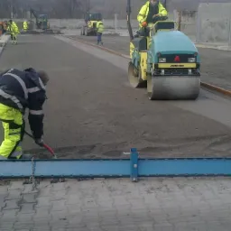 Walec Ammann ubija świeżo rozsypany tłuczeń na drodze, pracownik wyrównuje powierzchnię łopatą, w tle widoczne inne maszyny budowlane.